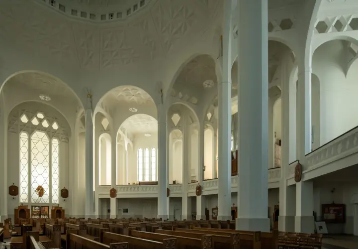 Jasne, nowoczesne wnętrze kościoła św. Rocha z rzędami drewnianych ławek i geometrycznymi zdobieniami sklepienia.