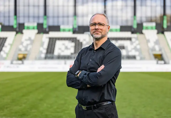 Rafał Smalec, w czarnej koszuli, stoi na tle stadionu piłkarskiego.