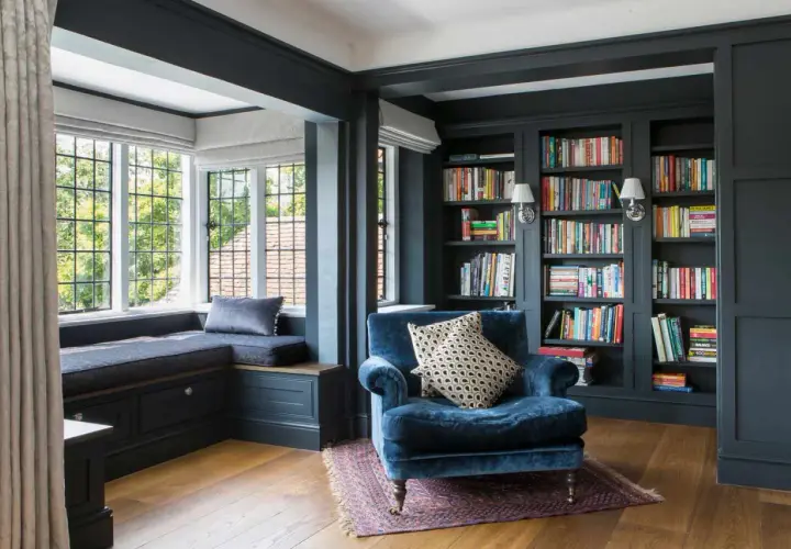 A plush blue armchair sits in a corner cozy reading nook, beside a window seat and floor-to-ceiling bookshelves filled with colorful books.