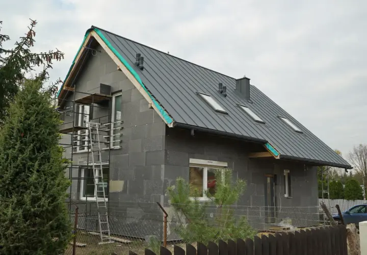 Rozbudowa starego domu w toku. Szary, nowoczesny budynek z metalowym dachem i oknami dachowymi, otoczony rusztowaniami.