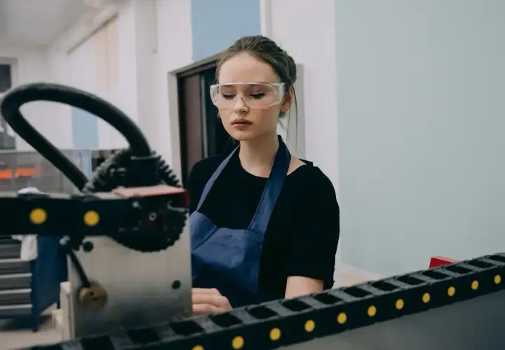 Młody pracownik produkcji w okularach ochronnych i fartuchu pracuje przy maszynie.