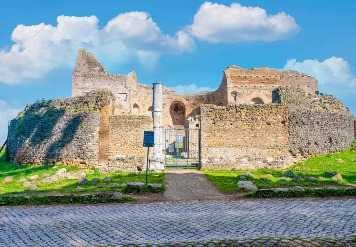 Ruines antiques sur l'Appia Antica à Rome, sous un ciel bleu parsemé de nuages blancs.