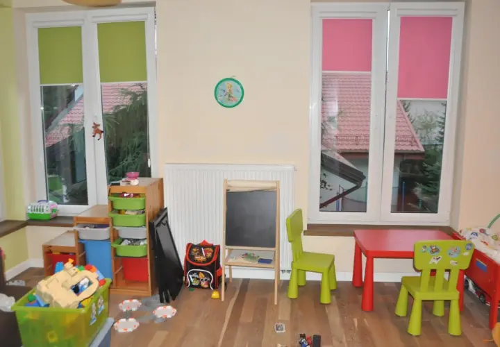 Pokój dziecięcy z zabawkami, stolikiem i krzesełkami. Okna z zielonymi i różowymi roletami, a za nimi widać dachy domów. Trofast inspiracje dla małych artystów.