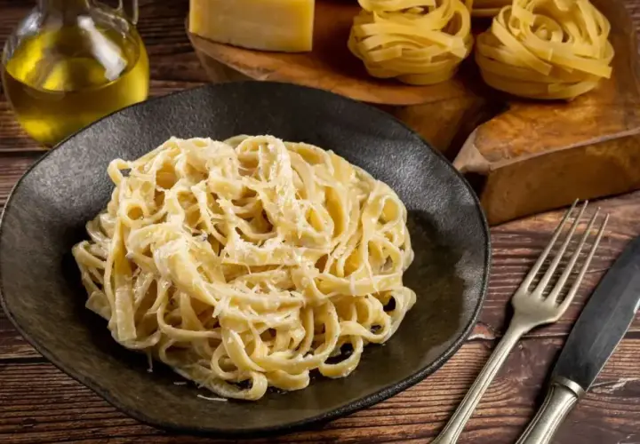 Talerz pełen tagliatelle z kremowym, oryginalnym sosem alfredo, obok butelka oliwy, ser i surowy makaron.