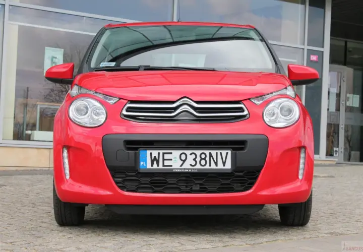 Czerwony Citroen C1 z tablicą rejestracyjną WE 938NV stoi przed budynkiem z dużymi oknami.