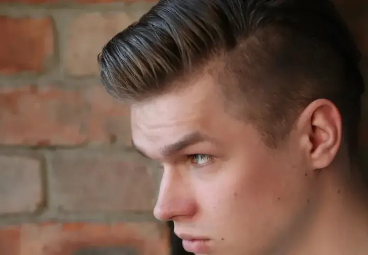 Joven con un corte de pelo moderno, un mid fade en pico, con el cabello peinado hacia arriba y a un lado, frente a una pared de ladrillos.