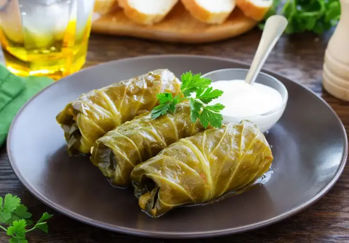 Tre involtini di cavolo cappuccio, guarniti con prezzemolo, serviti con una ciotolina di salsa bianca e un bicchiere di bevanda dorata.
