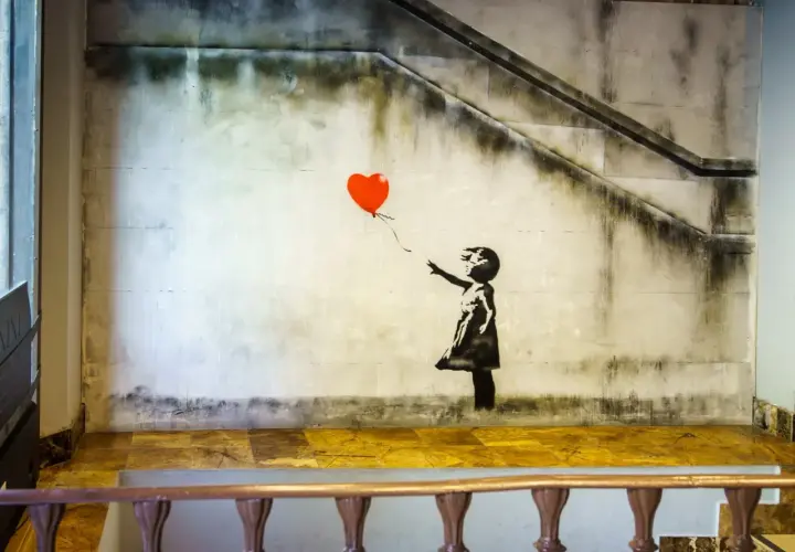 Dziewczyna z balonami, Banksy. Dziecko w czarnej sukience sięga po czerwony balon w kształcie serca na tle zniszczonej ściany ze schodami.