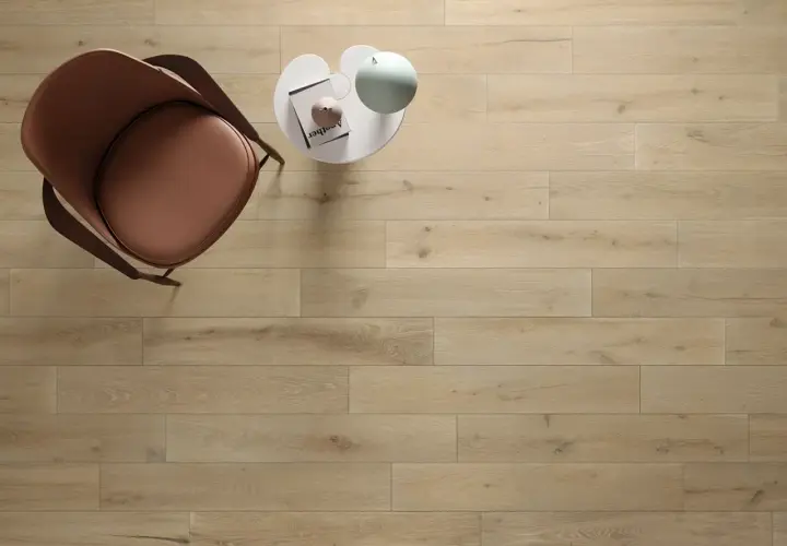 Un tavolino bianco con libro e lampada, accanto a una poltrona marrone, su un pavimento in legno con schemi posa a correre sfalsata.