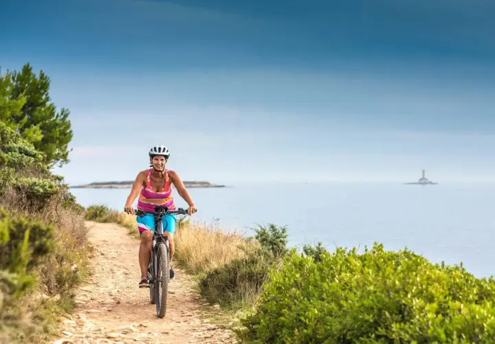 Woman enjoys cycling tours in Slovenia, biking along a coastal path with the sea and a distant lighthouse.