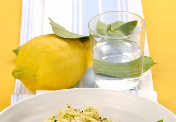 Un piatto di riso zucchine e limone, con un limone fresco e un bicchiere d'acqua.