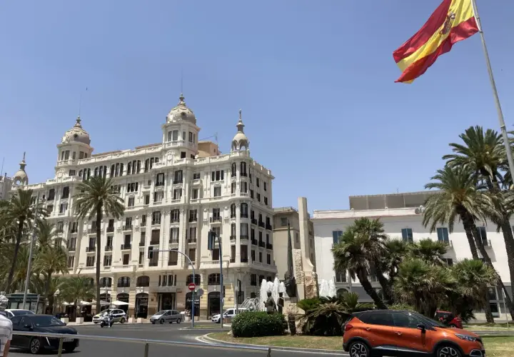 Alicante, pogoda marzec: słoneczny dzień, palmy i hiszpańska flaga nad białym budynkiem.