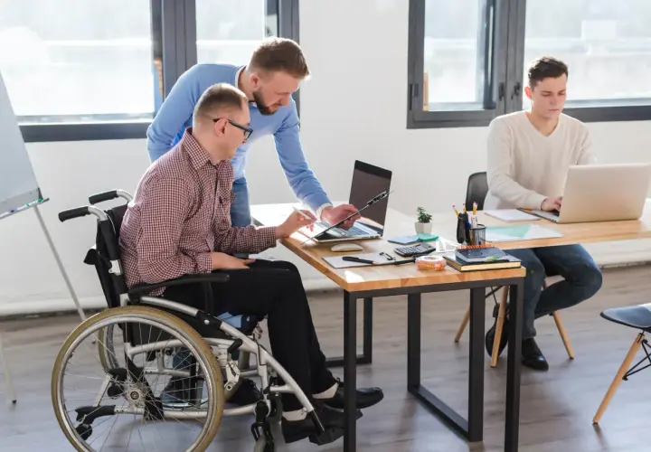 Dwóch mężczyzn pracuje przy biurku. Jeden siedzi na wózku inwalidzkim, drugi stoi obok. Trzeci mężczyzna pracuje przy laptopie przy innym biurku. To przykład efektywnego czasu pracy osoby niepełnosprawnej.