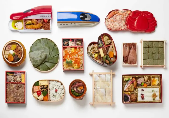 A variety of Japanese bento boxes are displayed, including train-shaped ones, a heart-shaped bento, and those filled with seafood, rice, and other delicacies.