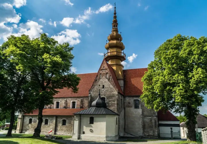 Kamienna świątynia z żółtą wieżą i czerwonym dachem, otoczona zielenią. W oddali widać fragmenty zabudowań, przypominające krajobraz wokół **zamku Krzyżtopór**.