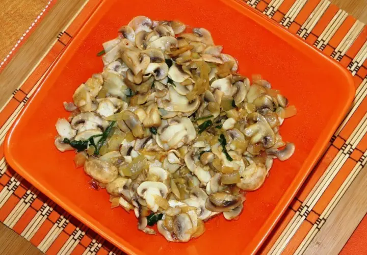 Champiñones salteados con cebolla y espinacas, servidos en un plato naranja sobre un mantel de bambú.