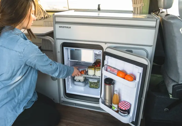 Une femme range des aliments dans un frigo pour van. Il contient des œufs, des légumes, des fruits et des boissons.