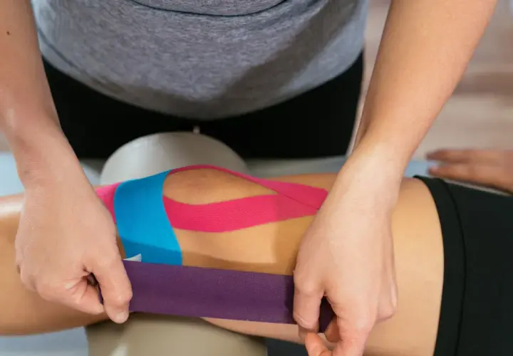 Fizjoterapeuta aplikuje kolorowe taśmy kinezyjne na kolano pacjenta, wspierając jego rehabilitację.