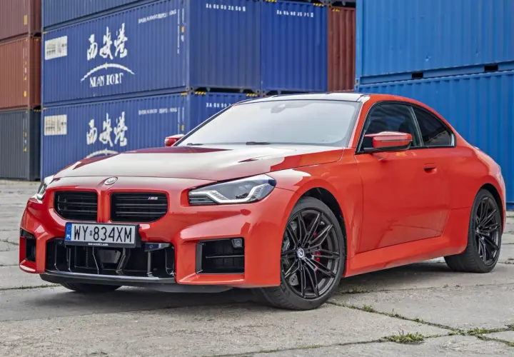 Pomarańczowe BMW M2 z czarnymi felgami, stojące przed niebieskimi kontenerami. Sprawdź aktualną bmw m2 cena!