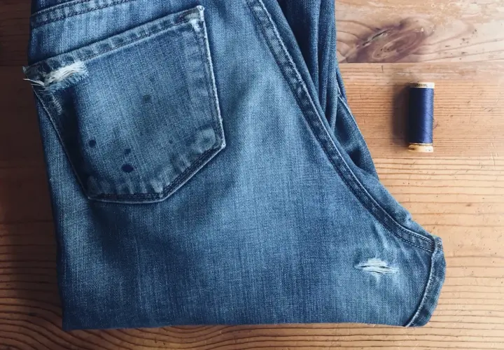 A pair of blue jeans with a small rip on the back pocket and a spool of navy thread, ready for how to patch jeans.