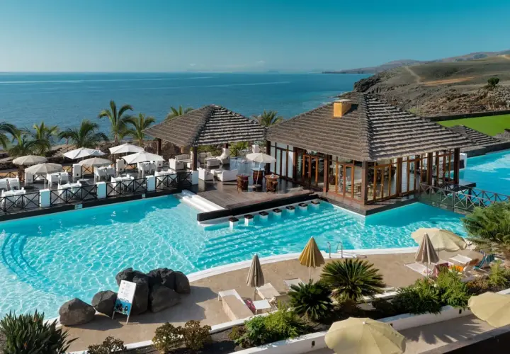 Un resort todo incluido con piscinas cristalinas, palmeras y vistas al mar. Mesas al aire libre y un bar acuático invitan a relajarse bajo el sol.