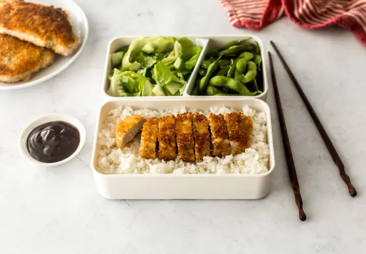 A delicious pork katsu bento box with rice, salad, edamame, and sauce.