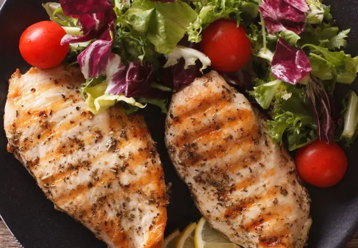 Due petti di pollo grigliati, conditi con erbe aromatiche, serviti con un'insalata fresca, pomodorini e spicchi di limone.