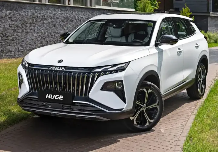 Biały, chiński SUV z imponującym grillem i napisem "HUGE" na masce, zaparkowany na podjeździe z kostki brukowej.