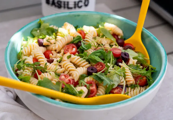 Ensalada de pasta espectacular con fusilli, tomates cherry, aceitunas negras y rúcula, servida en un cuenco azul con cucharas amarillas.