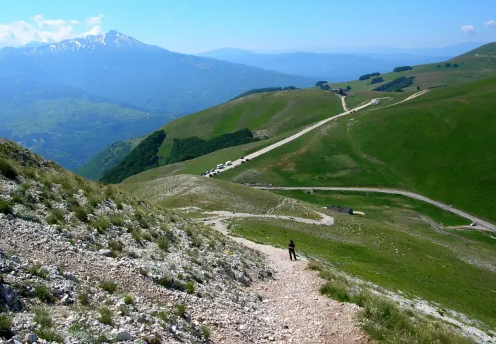 A person walks on a gravel path on a green mountain slope with a road winding through the hills and a snow-capped mountain in the distance.
