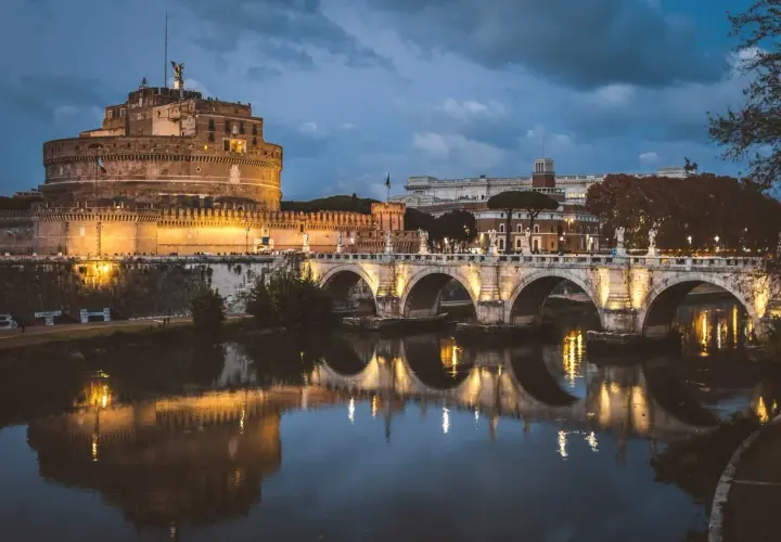 Le Castel Sant'Angelo, illuminé la nuit, se reflète dans le Tibre, avec un pont arqué en avant-plan.