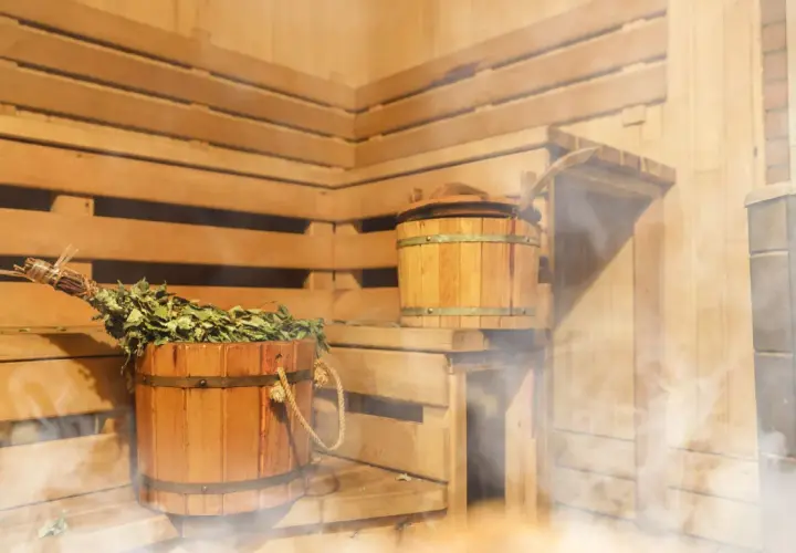Drewniana sauna z miotełką i kadzią, idealna na zakwasy po treningu. Para unosi się, tworząc relaksującą atmosferę.