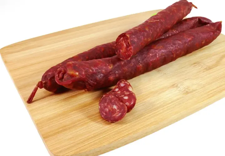 Trzy długie kiełbaski chorizo leżą na drewnianej desce. Jedna jest przekrojona, ukazując charakterystyczny, czerwony środek.