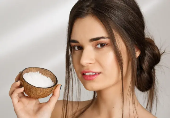Mujer sonríe sosteniendo una cáscara de coco con gránulos blancos, ideal para el aceite de coco para el pelo.