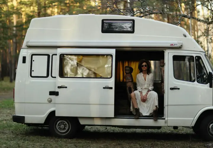 Kobieta w białej sukience siedzi w otwartych drzwiach białego kampera, otoczona lasem. Idealne polskie caravaning miejscówki na wakacje.