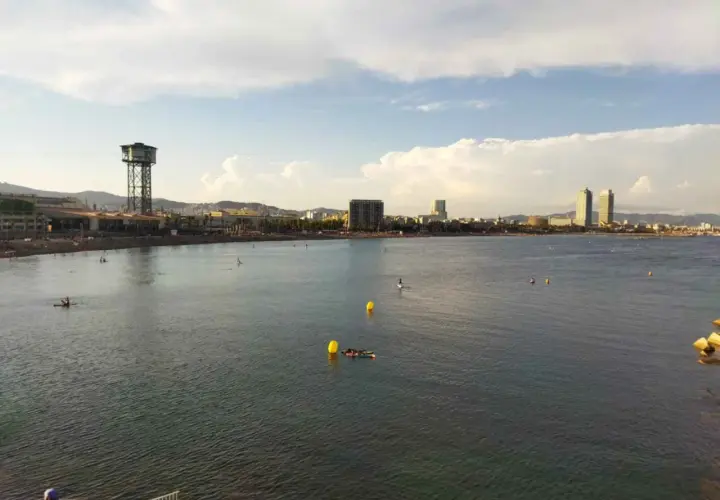 Vista de la playa y el mar con el icónico hotel vela al fondo. Personas disfrutan del agua en tablas de paddle surf.
