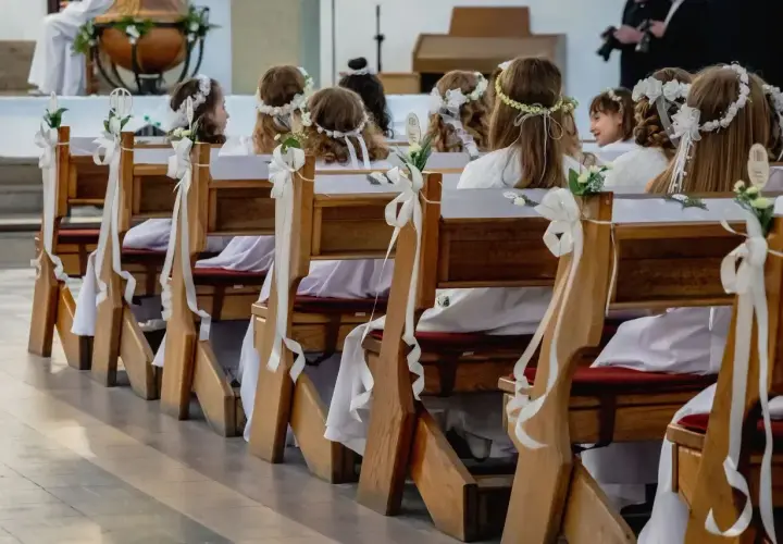Dzieci w bieli, z wiankami na głowach, siedzą w ławkach kościelnych. Czekają na ważny moment, zastanawiając się, ile trwa komunia.