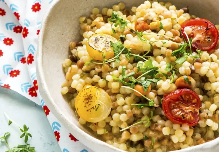 Un plato de pasta pequeña, que es semola, con tomates cherry asados y rodajas de limón, adornado con hierbas frescas.