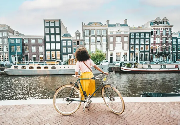 Kobieta z rowerem na tle amsterdamskich kamienic i kanału. Idealne miejsce na odkrywanie tras rowerowych Europa.