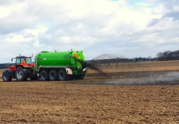Czerwony traktor z zielonym wozem asenizacyjnym rozprowadza poferment z biogazowni na polu.