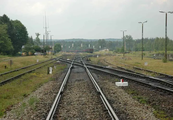 Rozstaw szyn kolejowych na stacji, z widocznymi zwrotnicami, budynkiem i drzewami w tle.