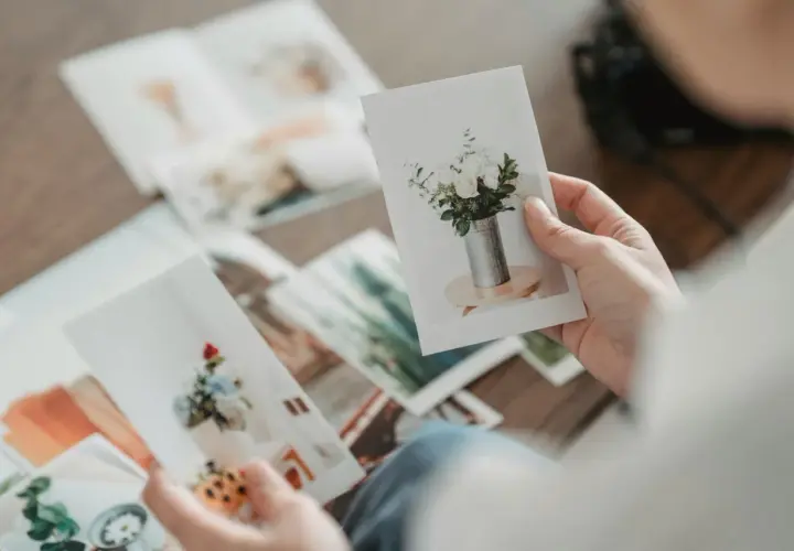 Dłonie przeglądają zdjęcia na papierze kredowym, ukazujące bukiety kwiatów w wazonach. To wspomnienia utrwalone na papierze fotograficznym.