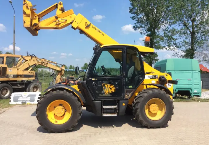 Żółty ładowarka teleskopowa JCB 531-70 Loadall stoi na placu. W tle koparka CAT i zielona kabina ciężarówki.