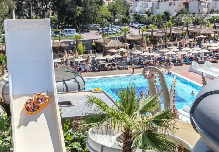 Dwie osoby zjeżdżają na pontonie ze zjeżdżalni wodnej w Atlantique Holiday Club. W tle basen i leżaki.