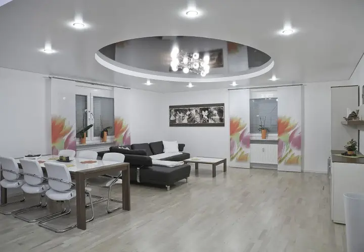 Nowoczesny salon z okrągłym sufitem napinanym, który odbija elegancki żyrandol. Wygodna kanapa i stół z krzesłami tworzą przytulną przestrzeń.