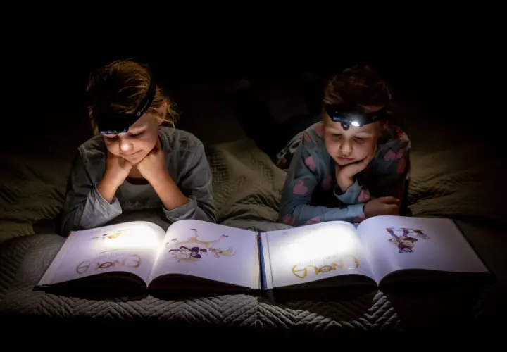 Dwie dziewczynki z latarkami na głowach czytają książkę w ciemności. To świetny sposób, by przygotować się na blackout i nie nudzić się!