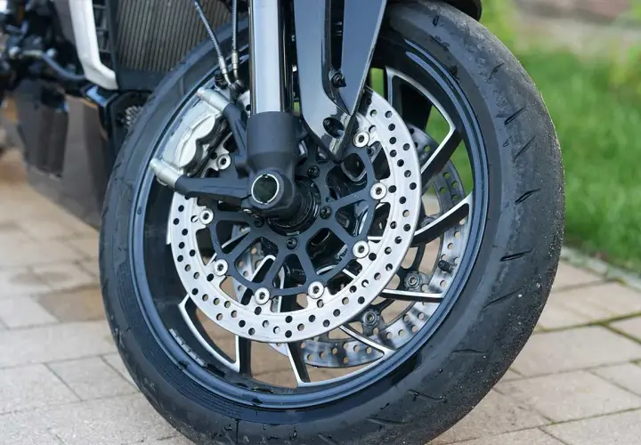 Primer plano de la rueda delantera de una motocicleta, con discos de freno perforados y pinzas. Se ve el neumático desgastado, listo para reparar rueda.