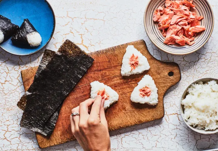 Making onigiri rice recipe: hands shaping rice balls with salmon filling, nori sheets, and bowls of rice and salmon.