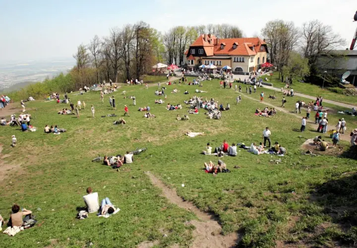Ludzie odpoczywają na trawie na zboczu góry. W tle budynek z czerwonym dachem. Widać ścieżki i drzewa.