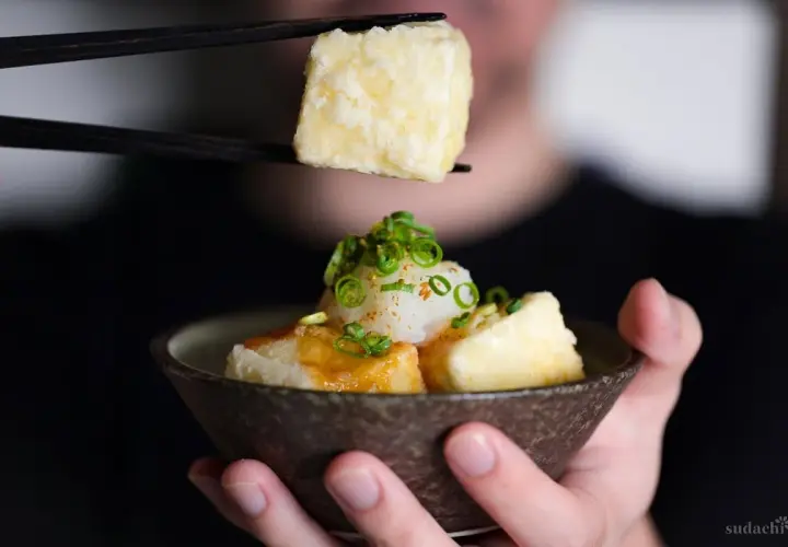 Chopsticks lift a piece of crispy age tofu, ready to be dipped into a savory sauce.